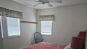 Ein Schlafzimmer mit Bett, Sessel, Schreibtisch, Deckenventilator und Fenster mit Jalousien.