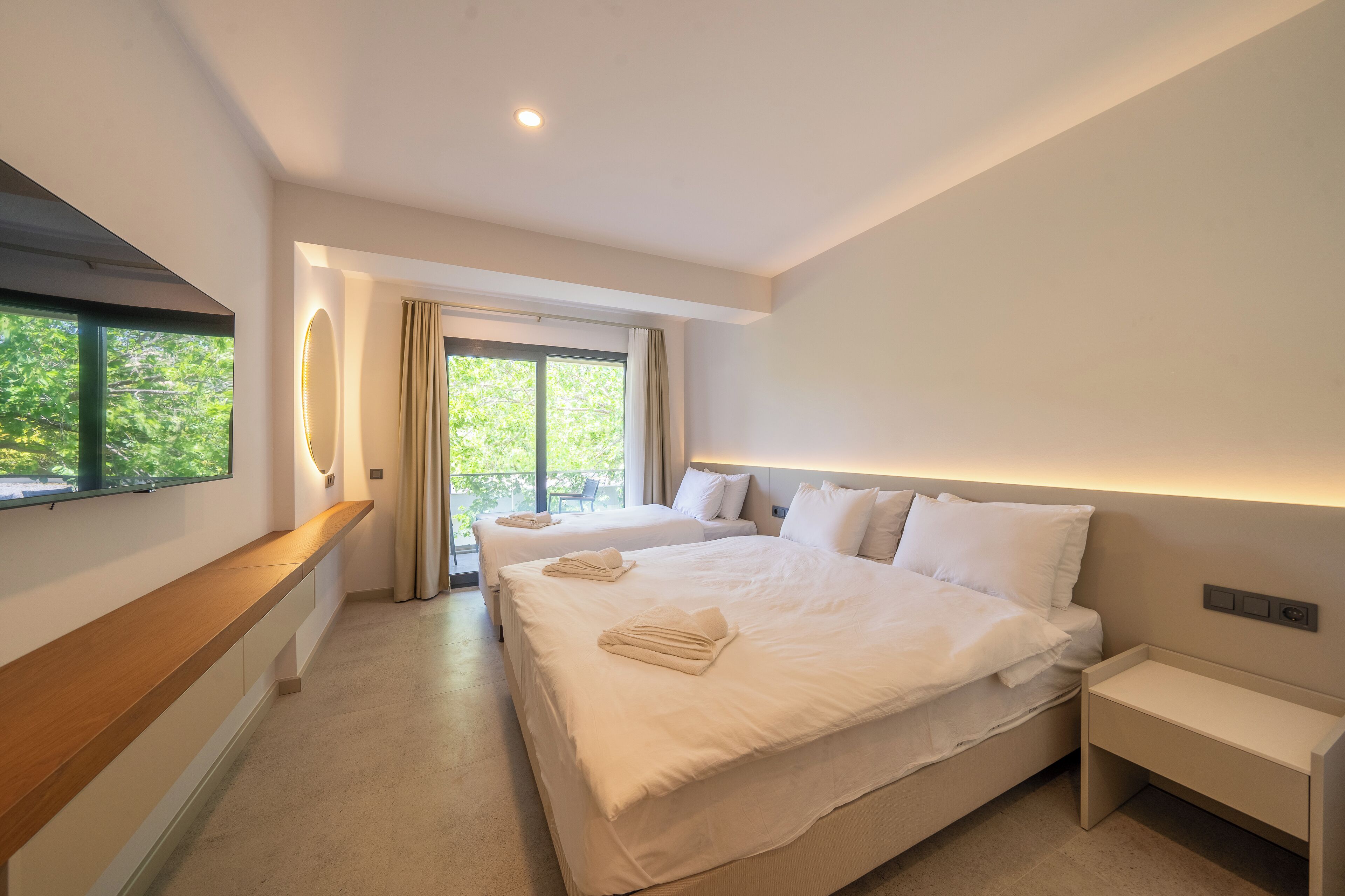 A modern hotel room with two beds, a wooden bench, a bedside table, and a large window with a view of greenery.