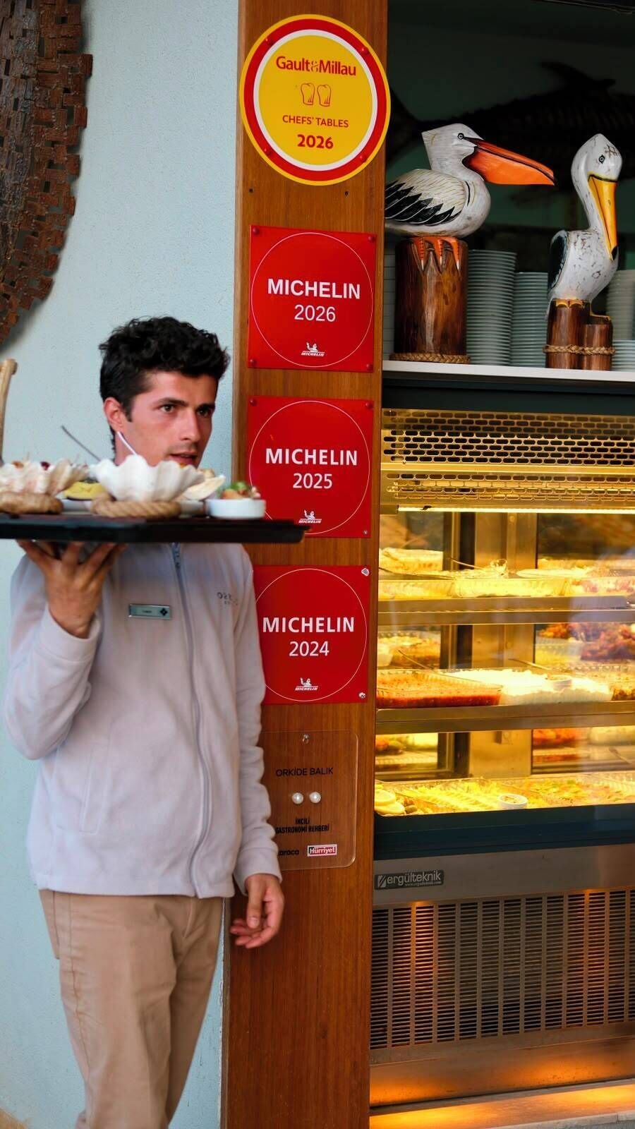 Une personne portant un plateau rempli de plats, une plaque commémorative récompensant un restaurant par un Guide Michelin, et une vitrine contenant des pâtisseries.