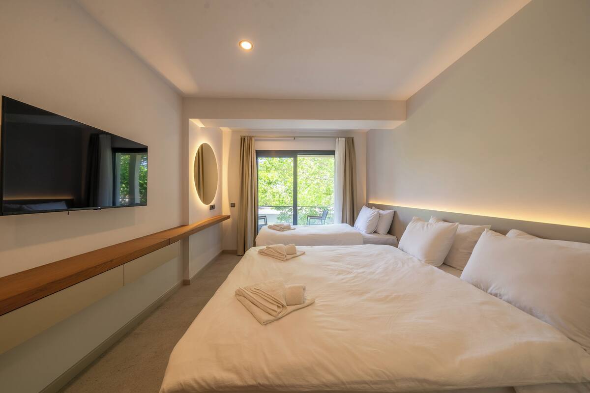 A hotel room with a large bed, a TV mounted on the wall, a balcony with a view of greenery, and a ceiling light.