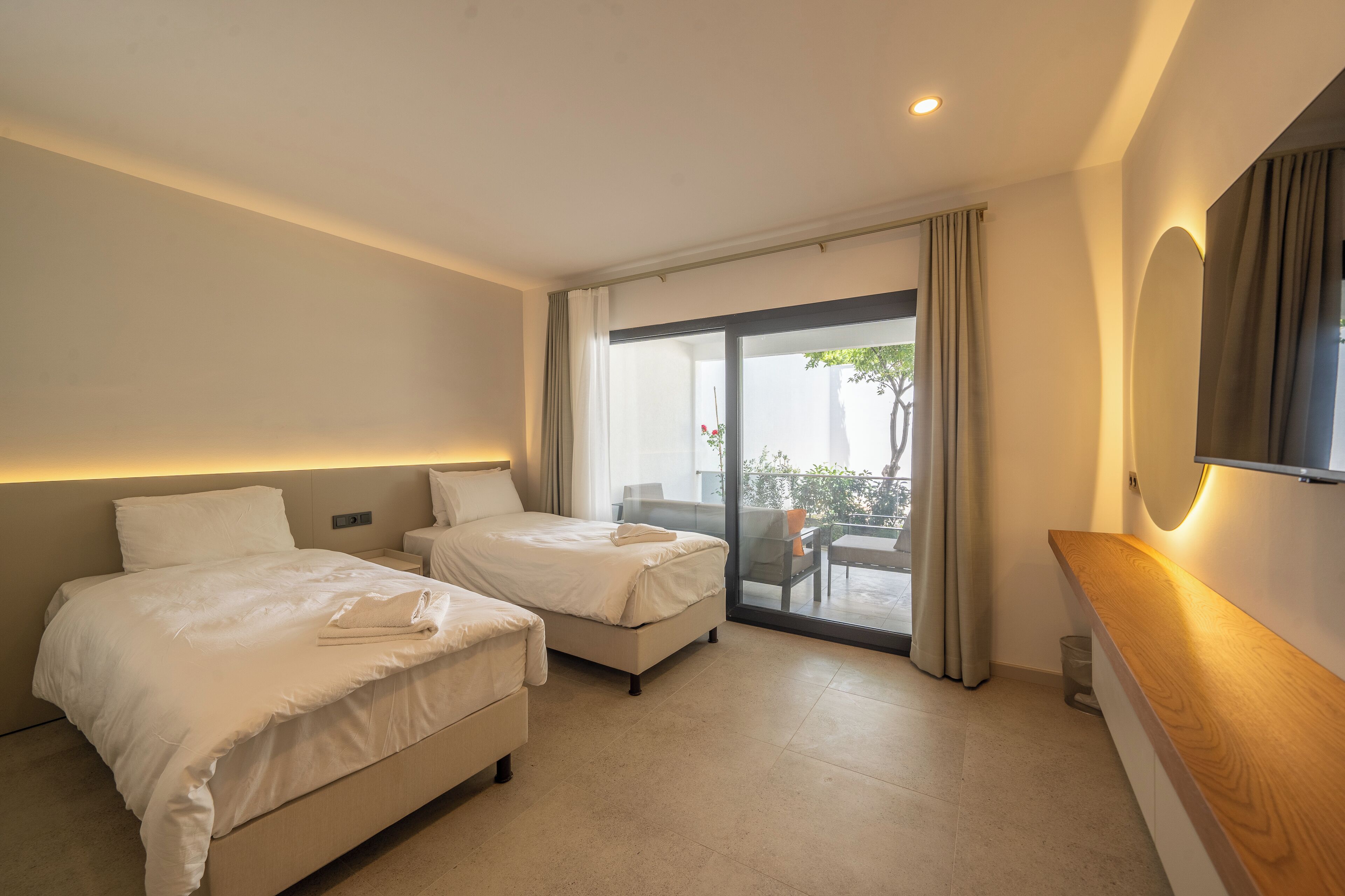 Habitación de hotel con dos camas, una mesita de noche de madera, un ventanal con cortinas y un balcón.