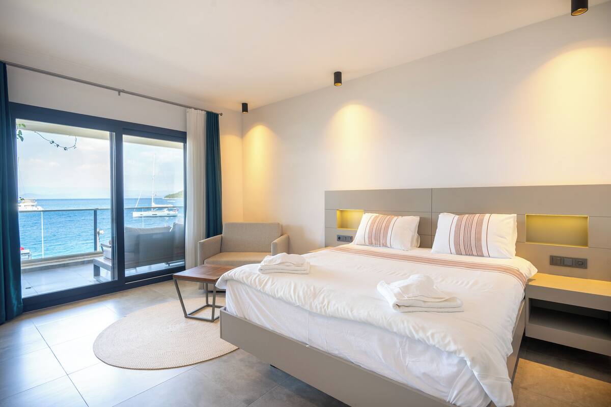 A modern hotel room with a large bed, a seating area, and a balcony with a view of the sea.