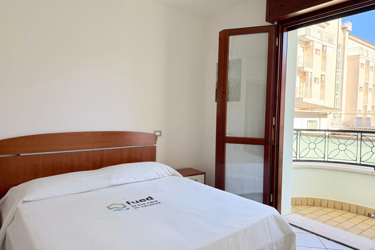 Ein Hotelzimmer mit Bett, Holz-Kopfteil, Nachttisch, einem Balkon mit Blick auf Gebäude und einer Tür mit Glasfenster.