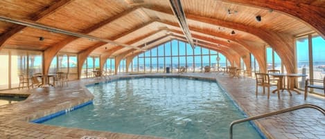 An indoor swimming pool with a wooden ceiling and brick flooring, surrounded by tables and chairs.