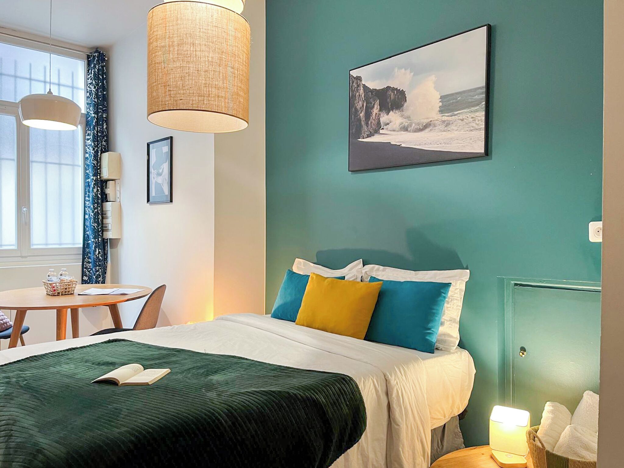 Una habitación con cama, una mesa y una silla. Hay un cuadro en la pared y una lámpara sobre la mesa.
