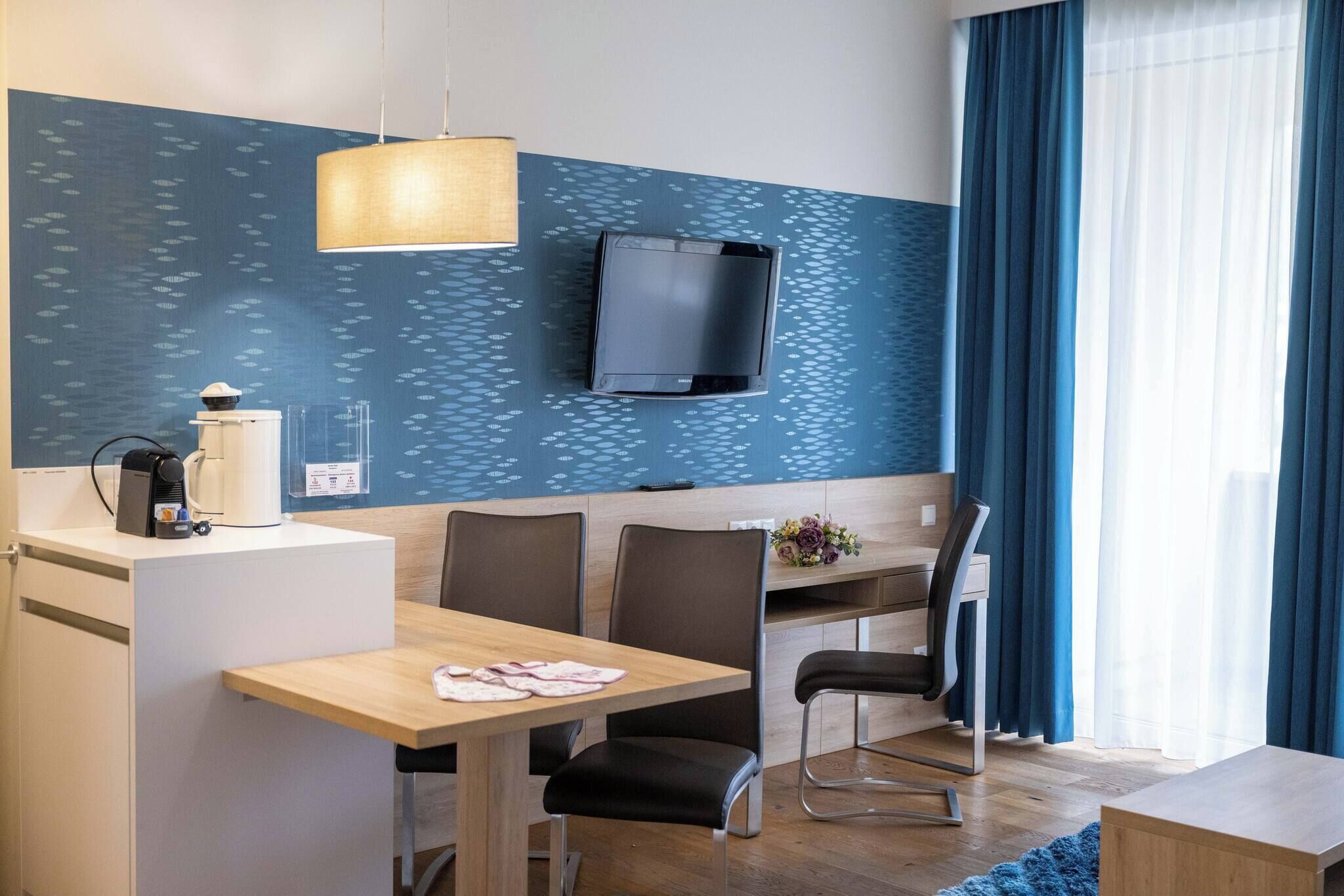A hotel room with a dining area, a TV mounted on the wall, and a window with curtains.