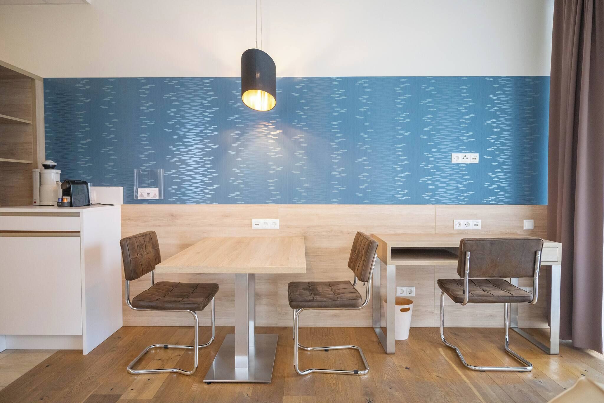A modern kitchen with a wooden table, two chairs, a hanging light, and a blue wall with a fish pattern.