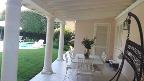 A covered patio with a pool, white columns, a table with chairs, and a hanging chair.