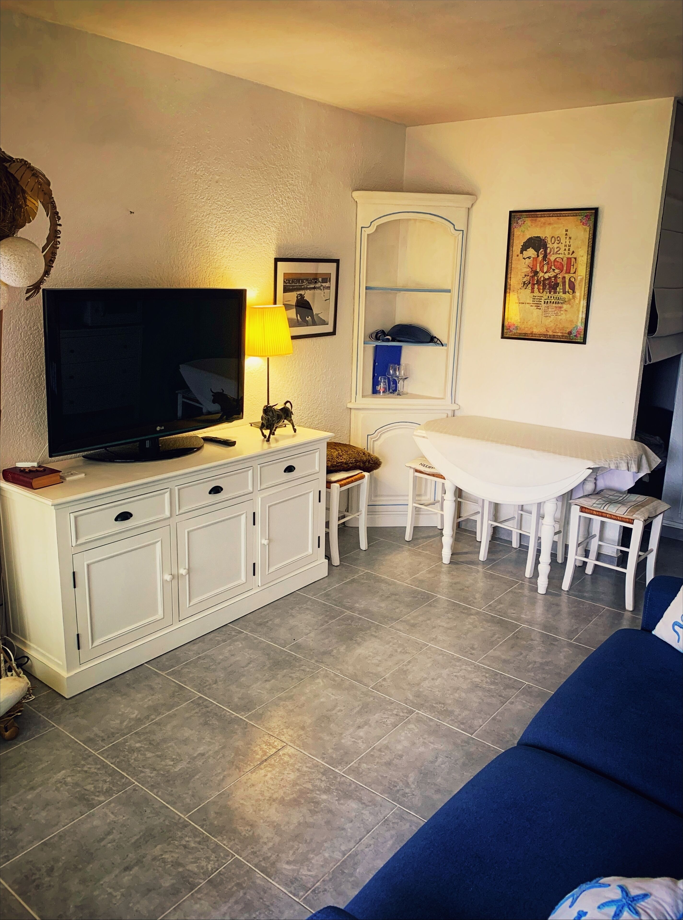 Una sala de estar con televisión, lámpara, mueble blanco, mesa de comedor con sillas y sofá azul.