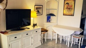 A living room with a TV, a lamp, a white cabinet, a dining table with chairs, and a blue sofa. - Studio by the sea with pool (Le Grau-du-Roi)
