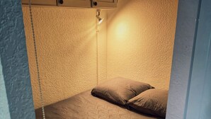A small, narrow room with a patterned bedspread, two pillows, and a single overhead light fixture. - Studio by the sea with pool (Le Grau-du-Roi)