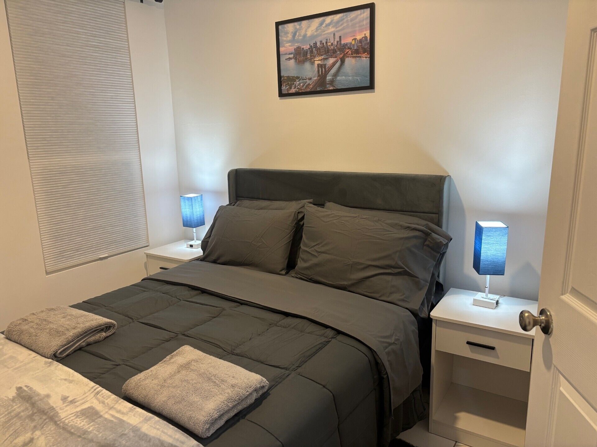 A neatly made bed with a dark-colored duvet and pillows, two bedside lamps, a window with blinds, and a framed picture on the wall.