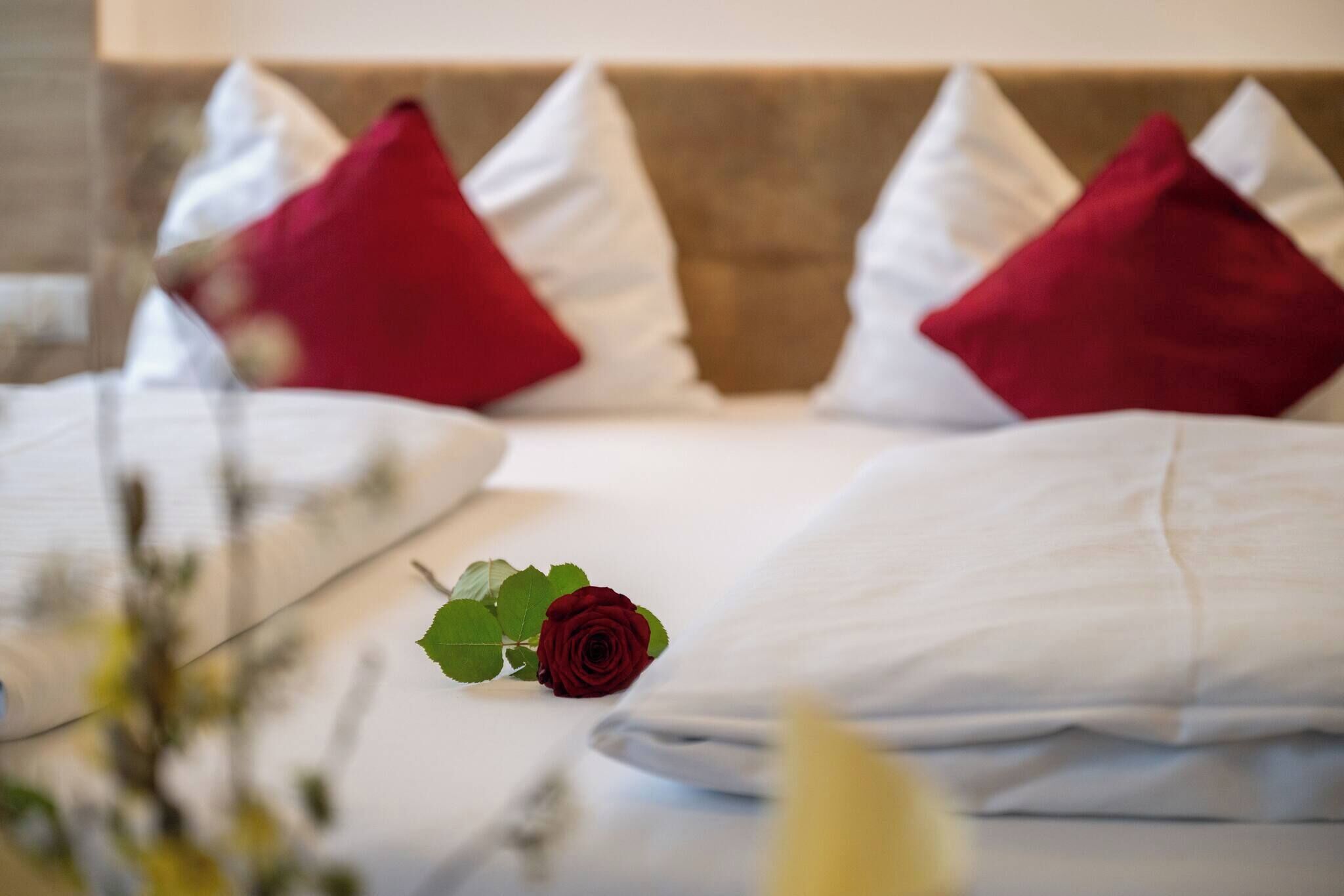 A neatly made bed with white and red pillows, a single red rose, and a blurred background suggesting a hotel room setting.