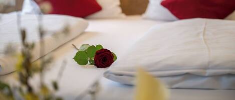 A neatly made bed with white and red pillows, a single red rose, and a blurred background suggesting a hotel room setting.