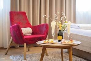 Ein modernes Zimmer mit einem roten Sessel, einem runden Holztisch mit zwei Tellern Essen und zwei Weingläsern, sowie einem Bett im Hintergrund.