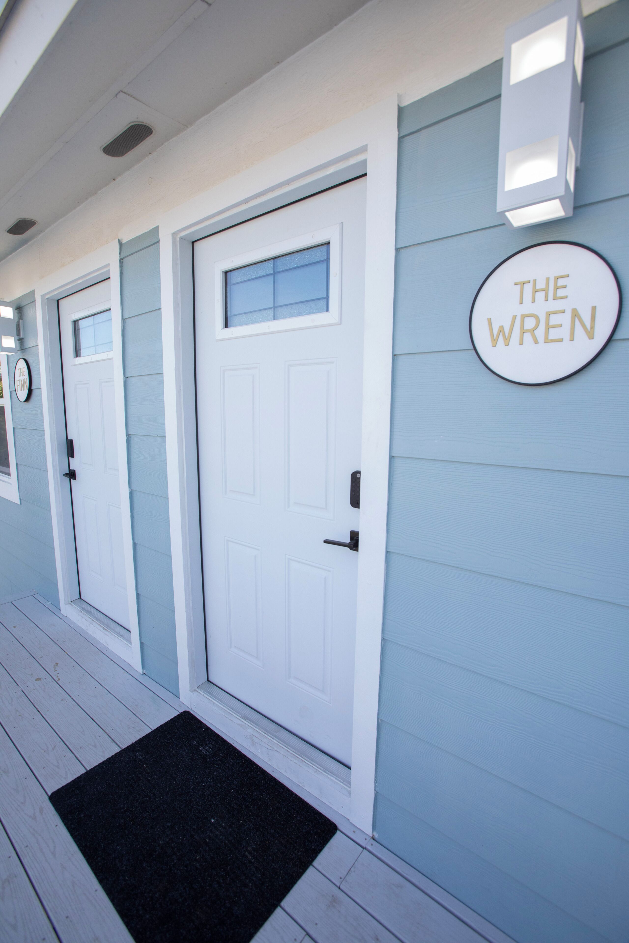 A building facade with three white doors, a black doormat, and a circular sign on the wall.
