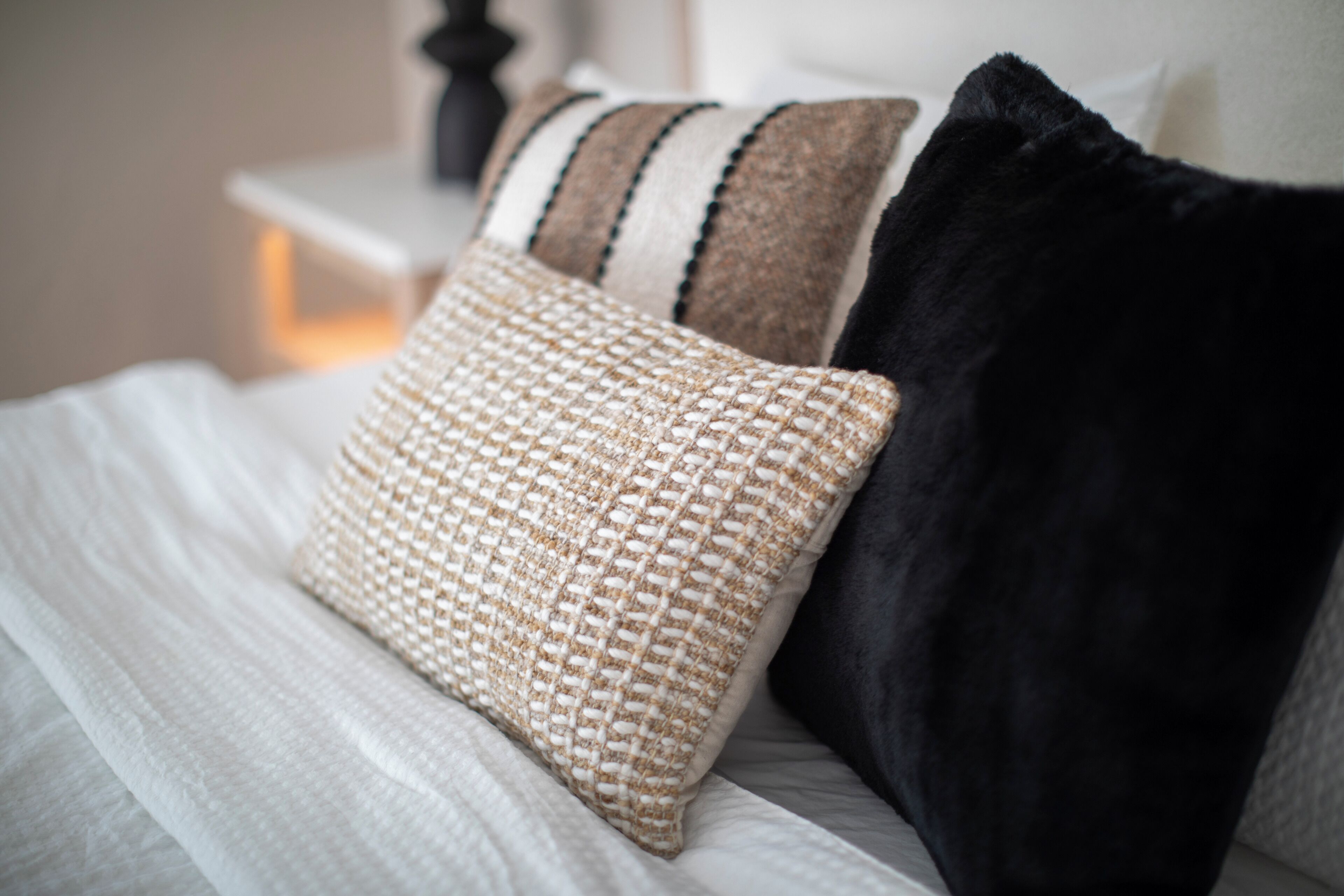 Ein Bett mit einer weißen Decke, einem beige gewebten Kissen und einem schwarzen Kissen.