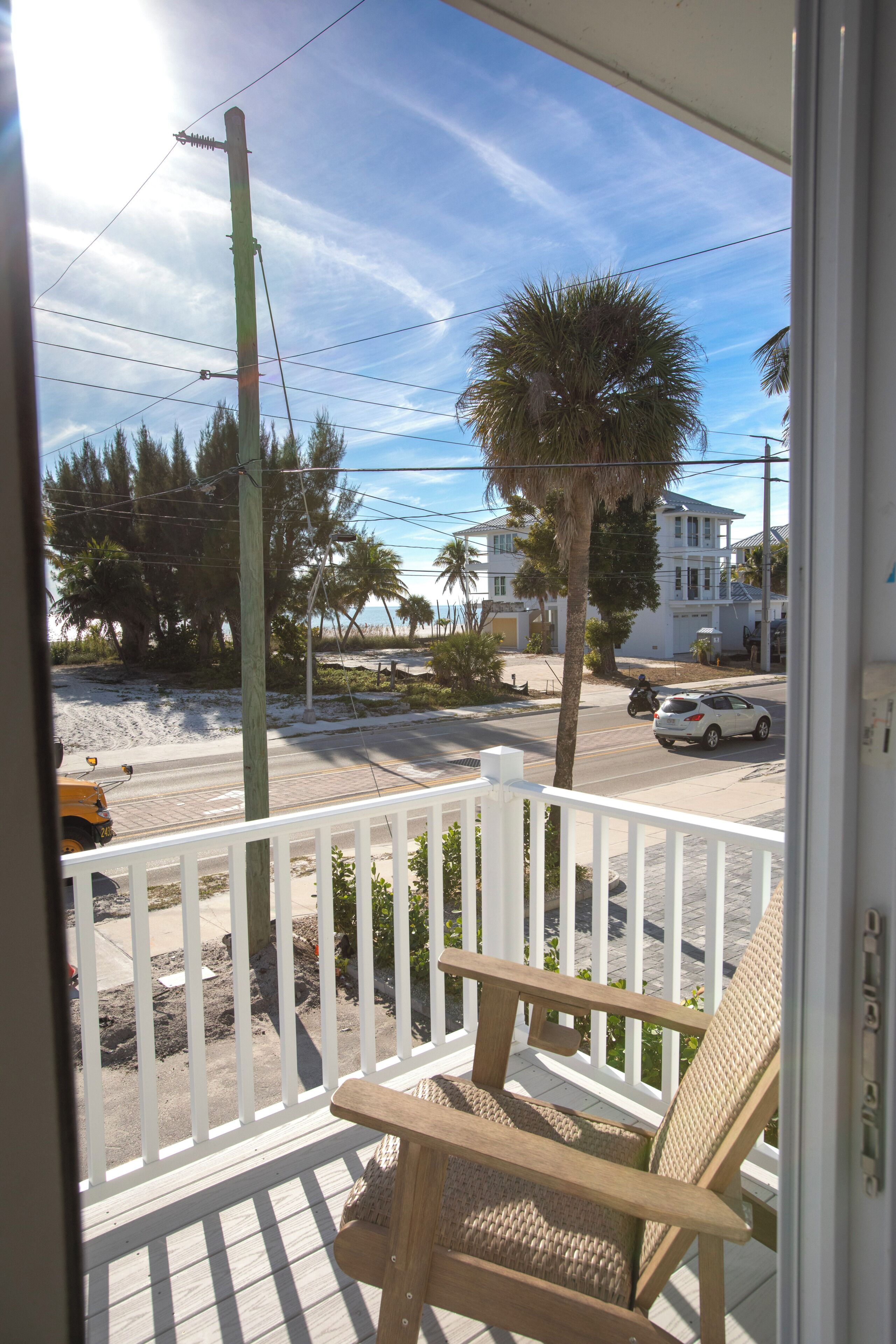 A balcony with a wooden chair, a white railing, a palm tree, and a view of a street with a white car and a building in the background.