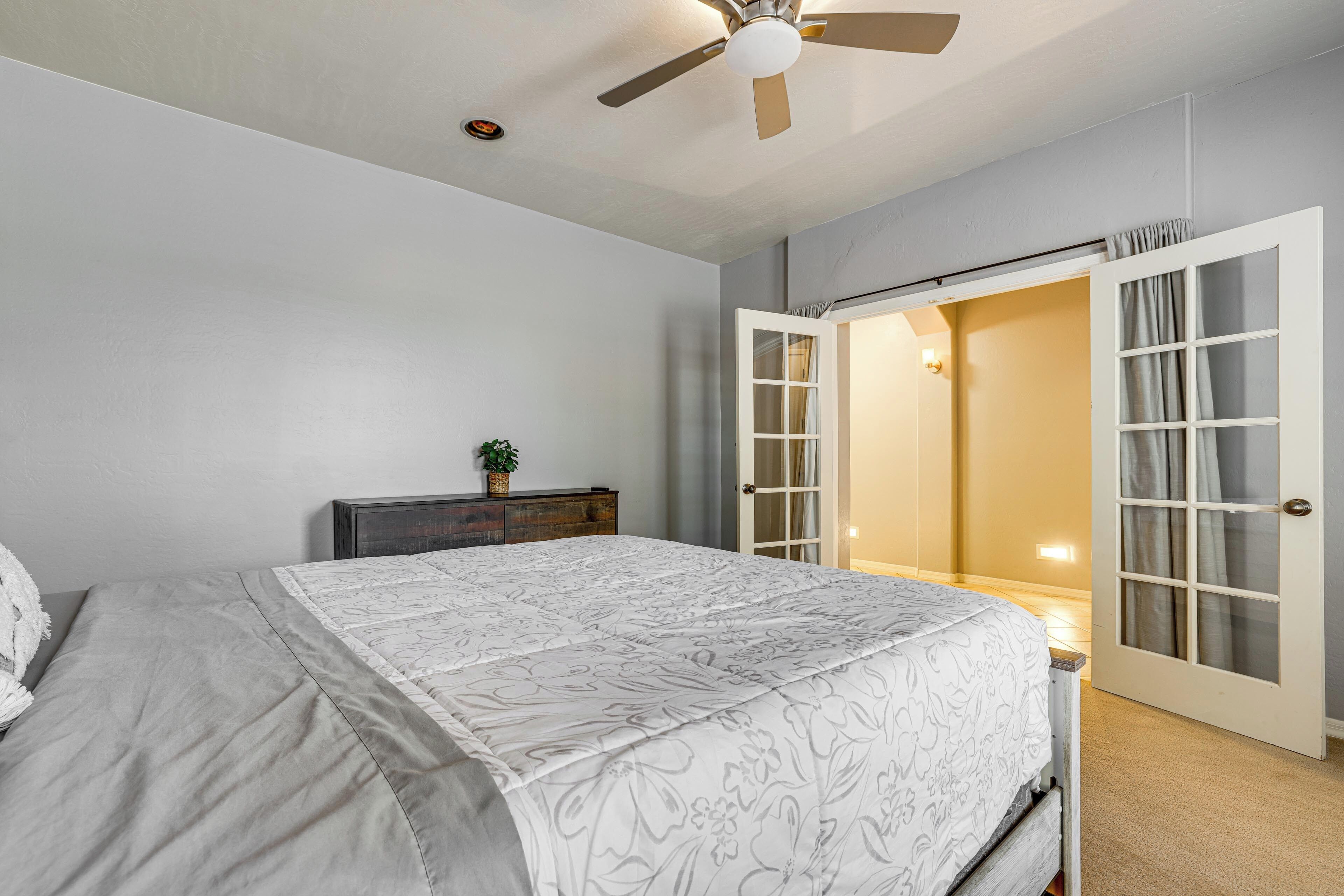 A bedroom with a bed, a ceiling fan, a nightstand, and a door leading to another room.