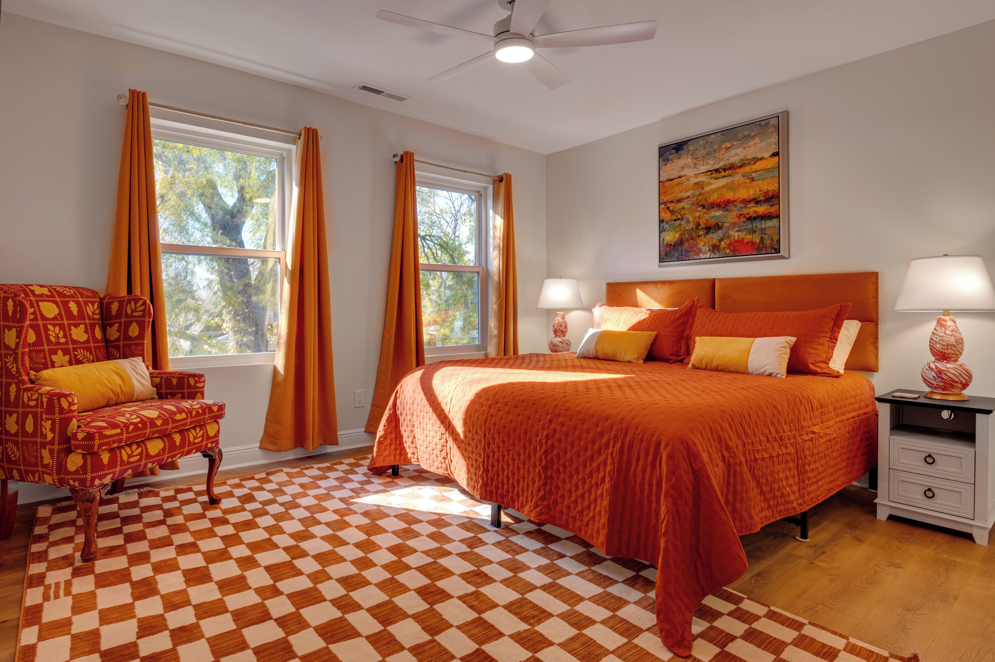 A bedroom with a checkered floor, a bed with orange bedding, a chair with a yellow cushion, and a painting on the wall.