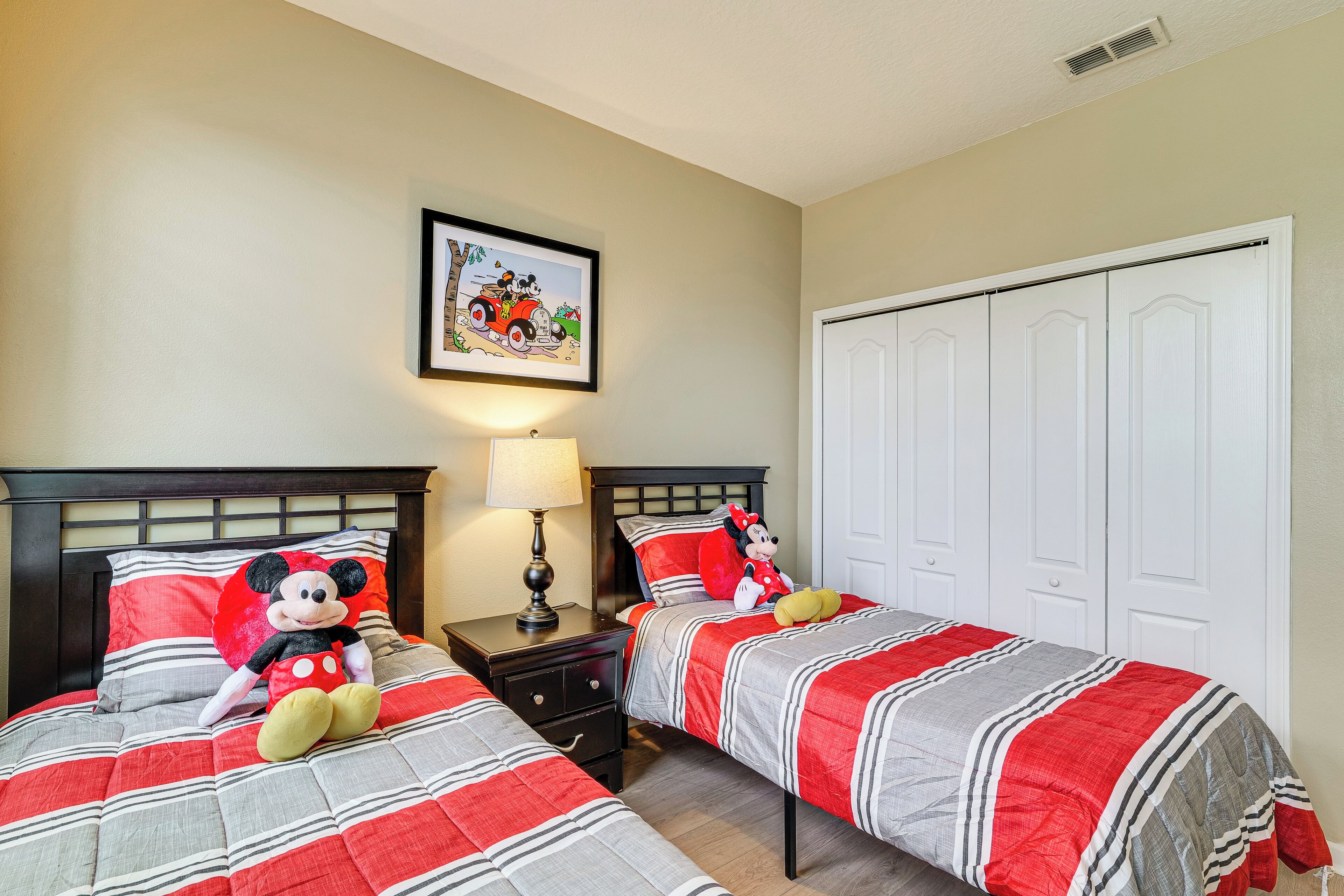 A hotel room with two beds, a nightstand with a lamp, a Mickey Mouse plush toy, and a framed picture on the wall.
