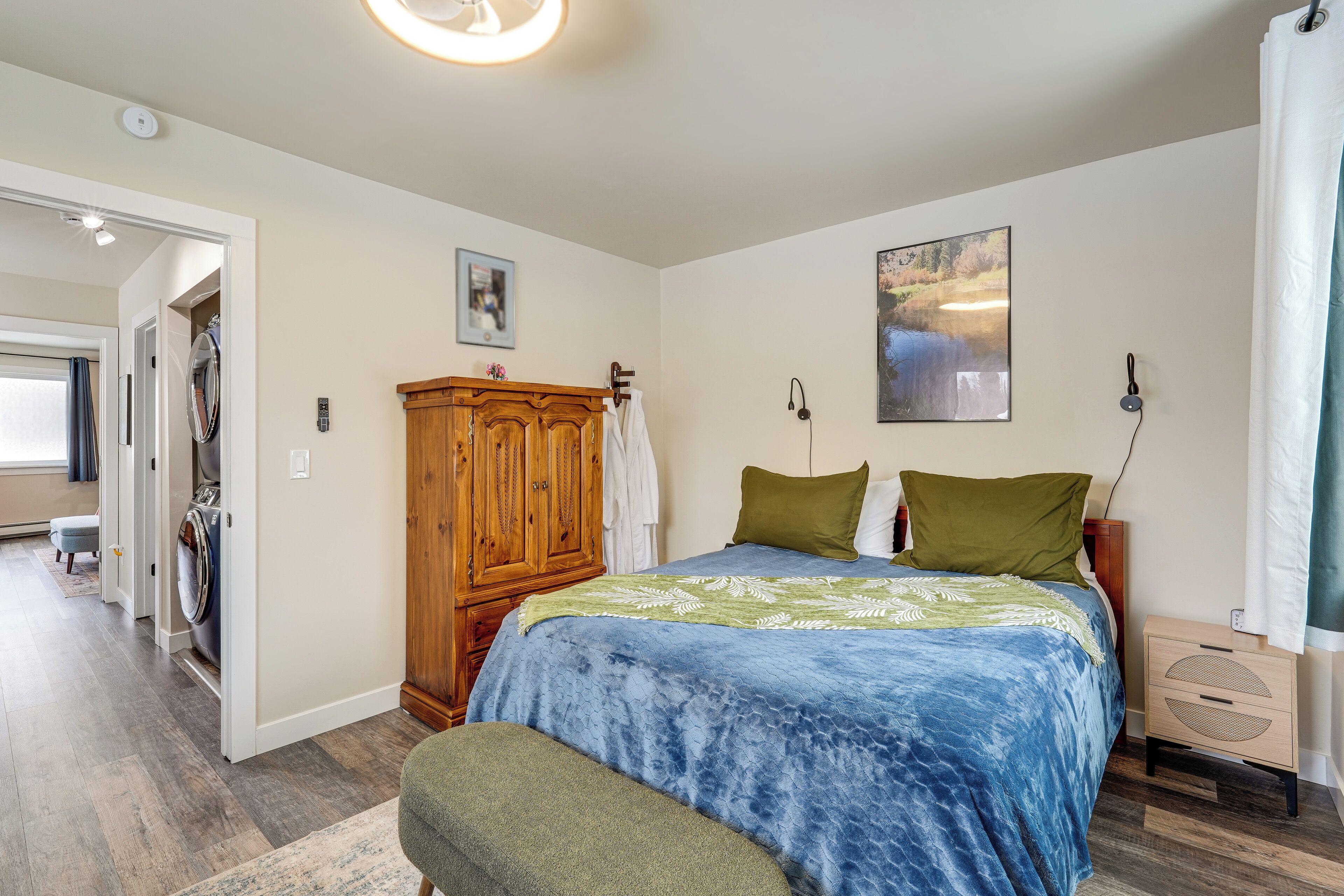 A bedroom with a bed, a wooden wardrobe, a washing machine, a bedside table, and a wall-mounted speaker.