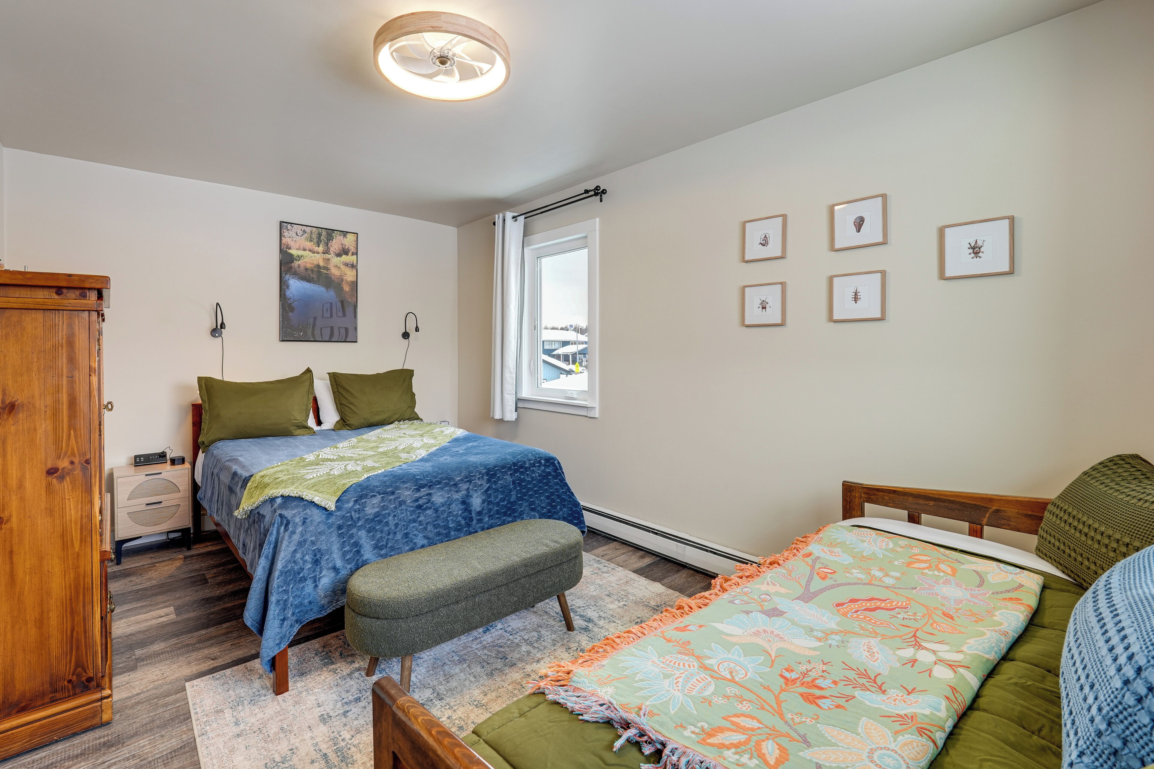 A bedroom with two beds, a wooden wardrobe, a window with a view, and framed pictures on the wall.