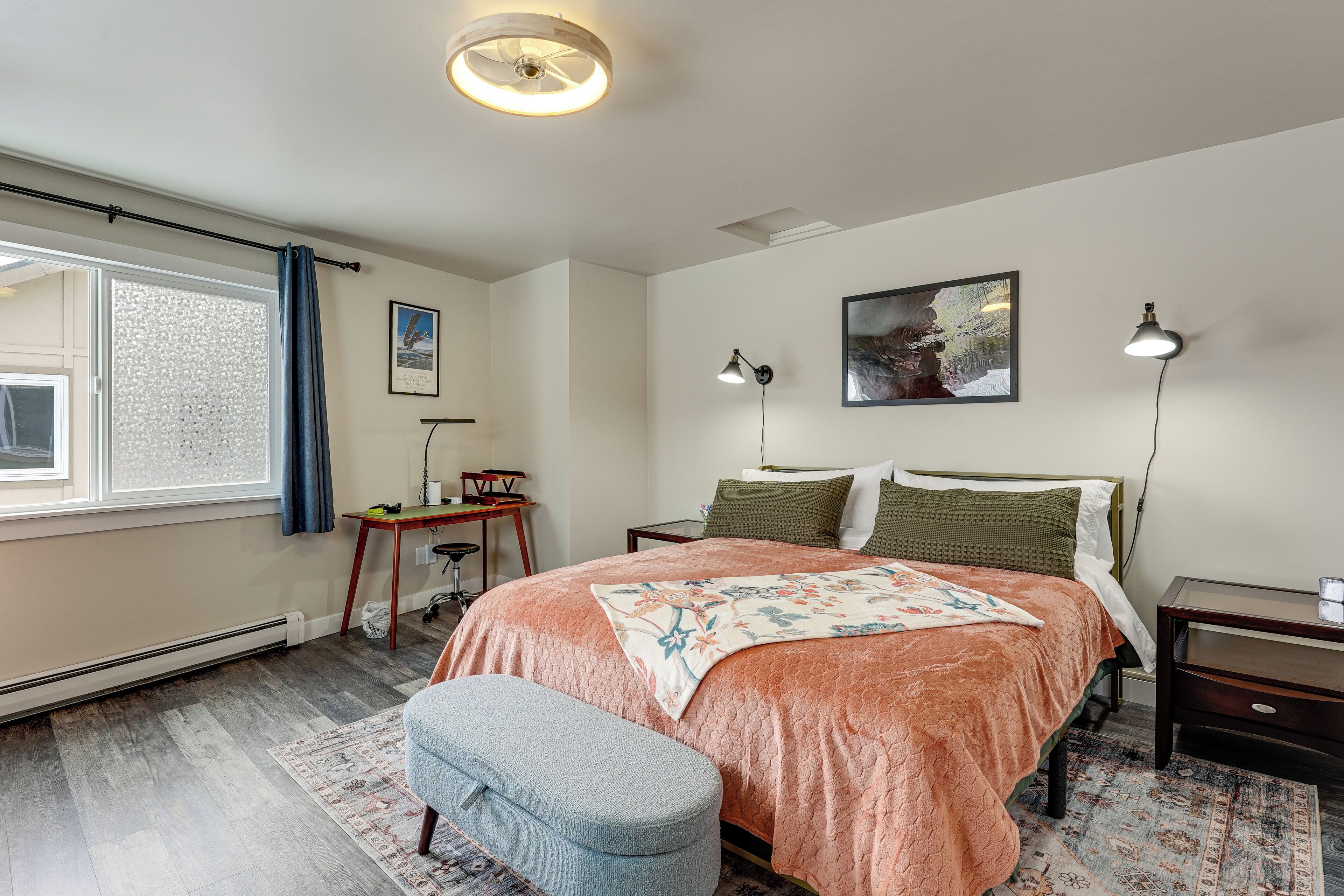 Ein Schlafzimmer mit Bett, Nachttischen, Schreibtisch, Stuhl, Fenster mit Vorhängen und Heizkörper.