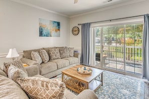 A living room with a sofa, a coffee table, a lamp, a clock, and a large window with a view of a garden. - Fall Golf Favorite 1st Floor Near CCU & Events (Myrtle Beach)