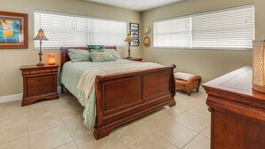 Une chambre à coucher avec un lit en bois, des tables de chevet, une commode, une lampe, un objet décoratif et une fenêtre avec des stores.