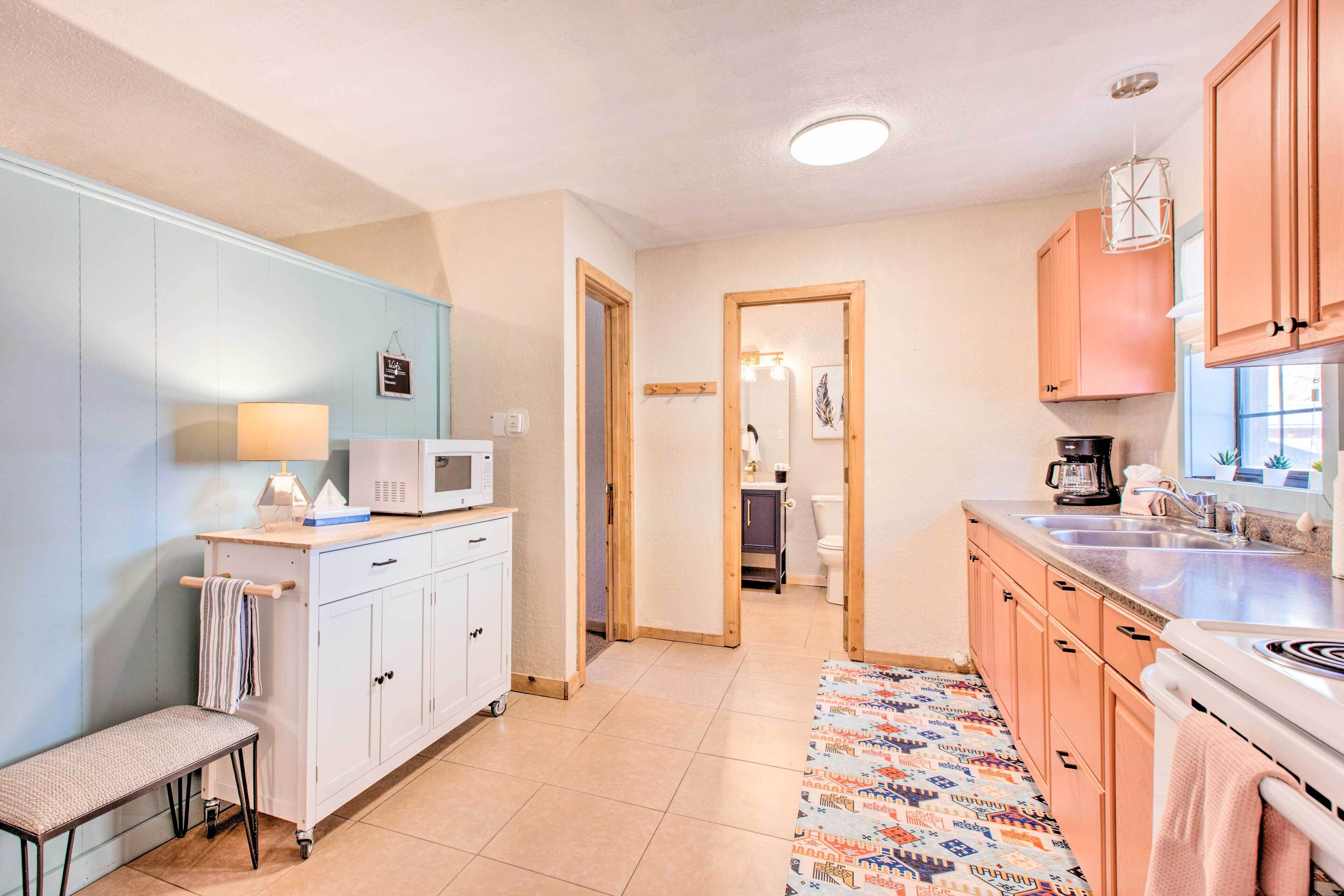 A compact kitchen with a microwave, a coffee maker, and a sink. There is a bench and a patterned rug.