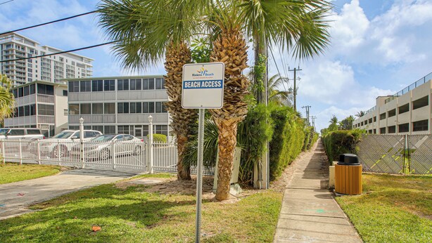Un panneau indiquant l’accès à la plage, un trottoir, des voitures garées et un bâtiment en arrière-plan.