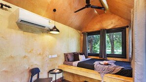 Ein kleines Zimmer mit Bett, Sessel, Tisch, Deckenventilator und einem fenster mit Vorhängen.