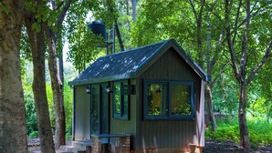 Eine kleine Holzhütte mit geneigtem Dach, großen Fenstern und einer Veranda mit Treppen, umgeben von Bäumen.