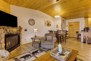 A living room with a stone fireplace, a television, a clock, and a dining area with wooden furniture.