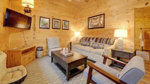 A cozy log cabin interior with a sofa, armchair, coffee table, and a flat-screen TV mounted on the wall. - Cozy Cabin By Gsmnp! Soaking Tub, Views + Fire Pit (Cosby)
