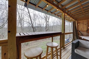Una terraza de madera con barandilla, dos taburetes, un sofá de mimbre y una hamaca.