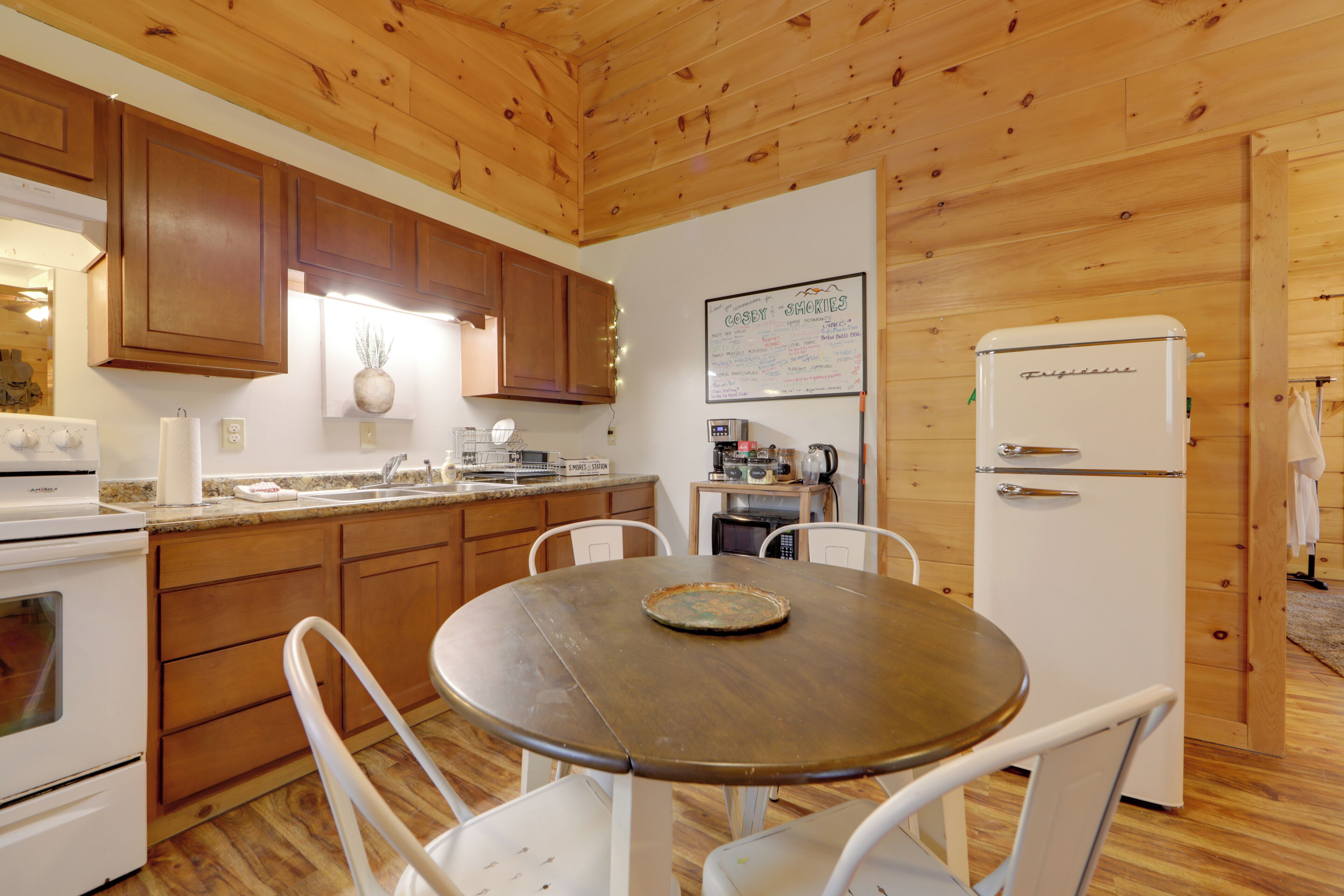 Una cocina con armarios de madera, un refrigerador blanco, una mesa de comedor con sillas y una pizarra en la pared.