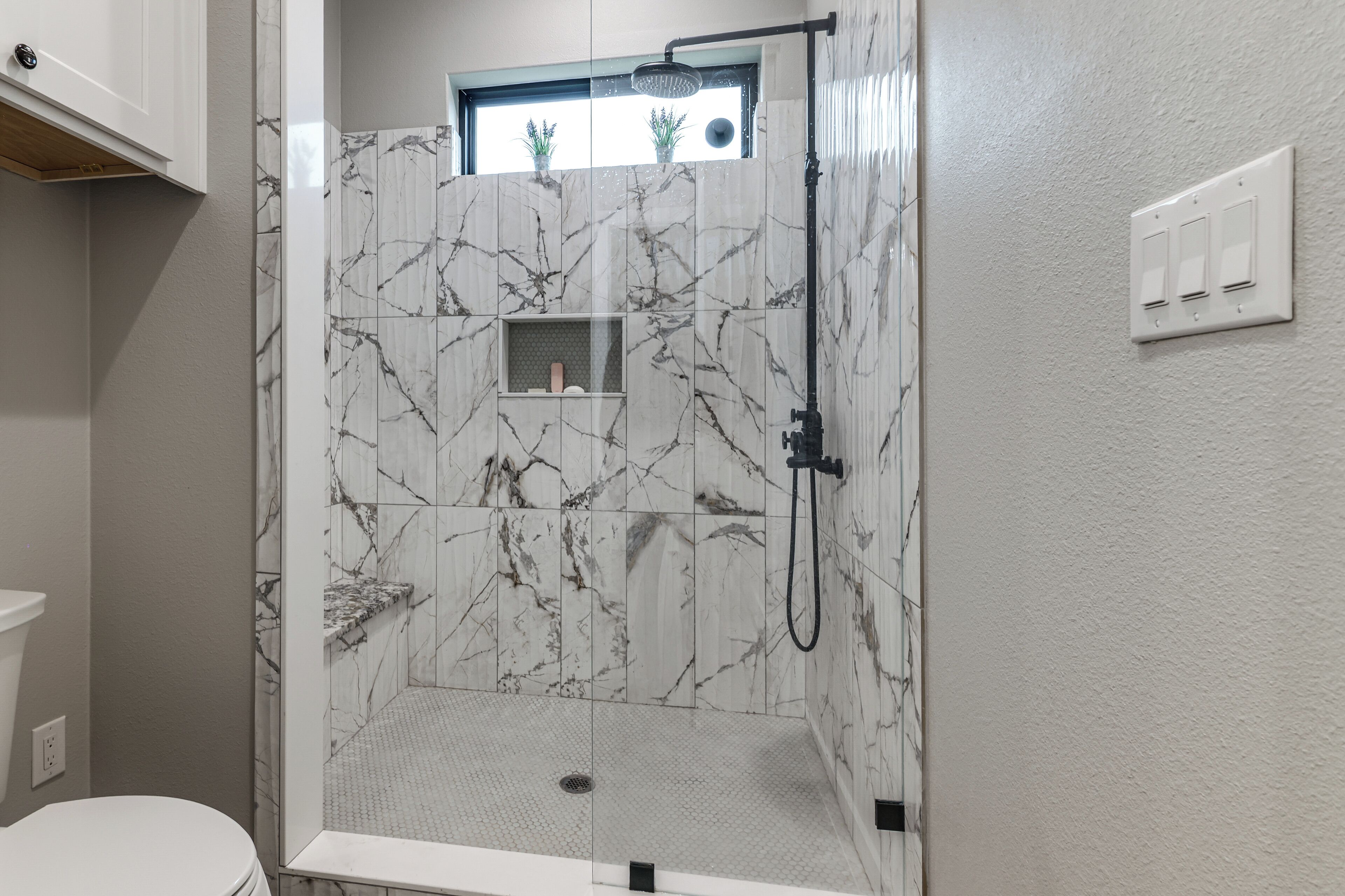 Eine barrierefreie Dusche mit Marmorwänden, einem Fenster mit Pflanzen und einer sichtbaren Toilette.