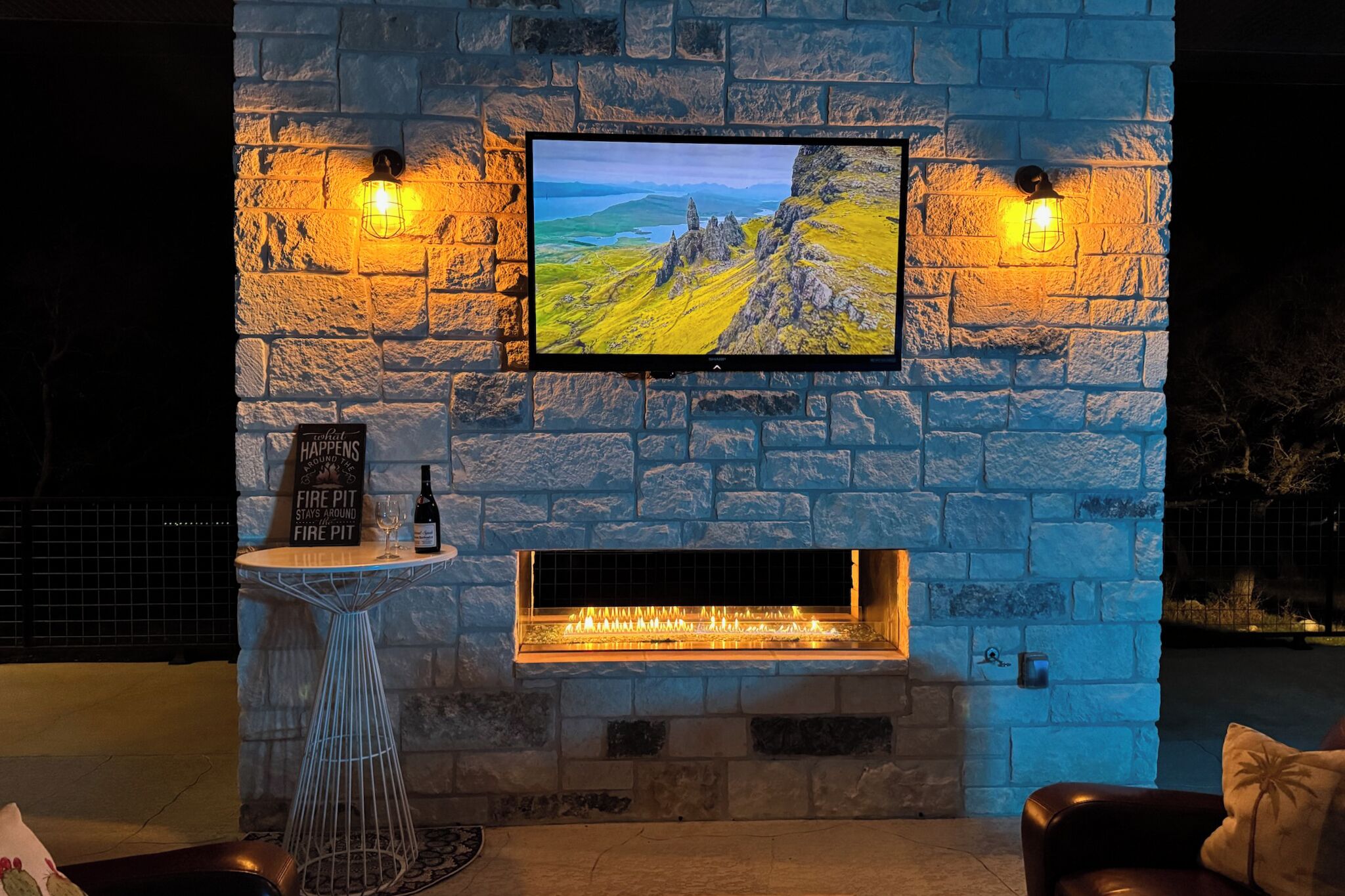 Een stenen open haard met een tv erboven, een klein tafeltje met een fles en glas, en een bordje met de tekst 'Fire Pit'.