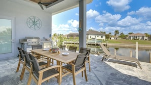 An outdoor patio with a wooden table and chairs, a grill, and a view of a lake and houses in the background. - Luxe Canal-front Getaway w/ Pool in Punta Gorda! (Punta Gorda)
