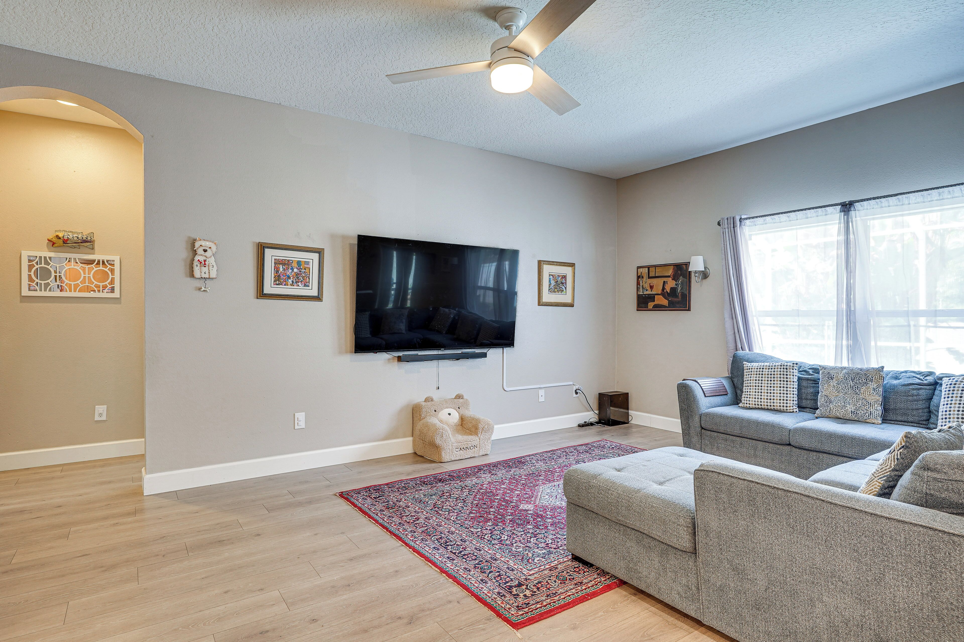 A living room with a flat-screen TV, grey sofa, red rug, and a ceiling fan.