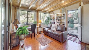A spacious living room with a wooden ceiling, a leather sofa, a bookshelf, and a large window. - Group Retreat on Flower Farm Near Sedona Trails (Cornville)