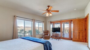 Une chambre à coucher avec un lit, un ventilateur de plafond, une télévision, une chaise et de grandes fenêtres donnant sur un beau paysage.