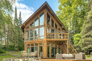 A wooden cabin with large windows, a balcony, and a staircase leading to an upper level.