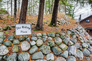 A sign reading "The Hiding Place" in a forested area with a stone wall and wooden cabin in the background.
