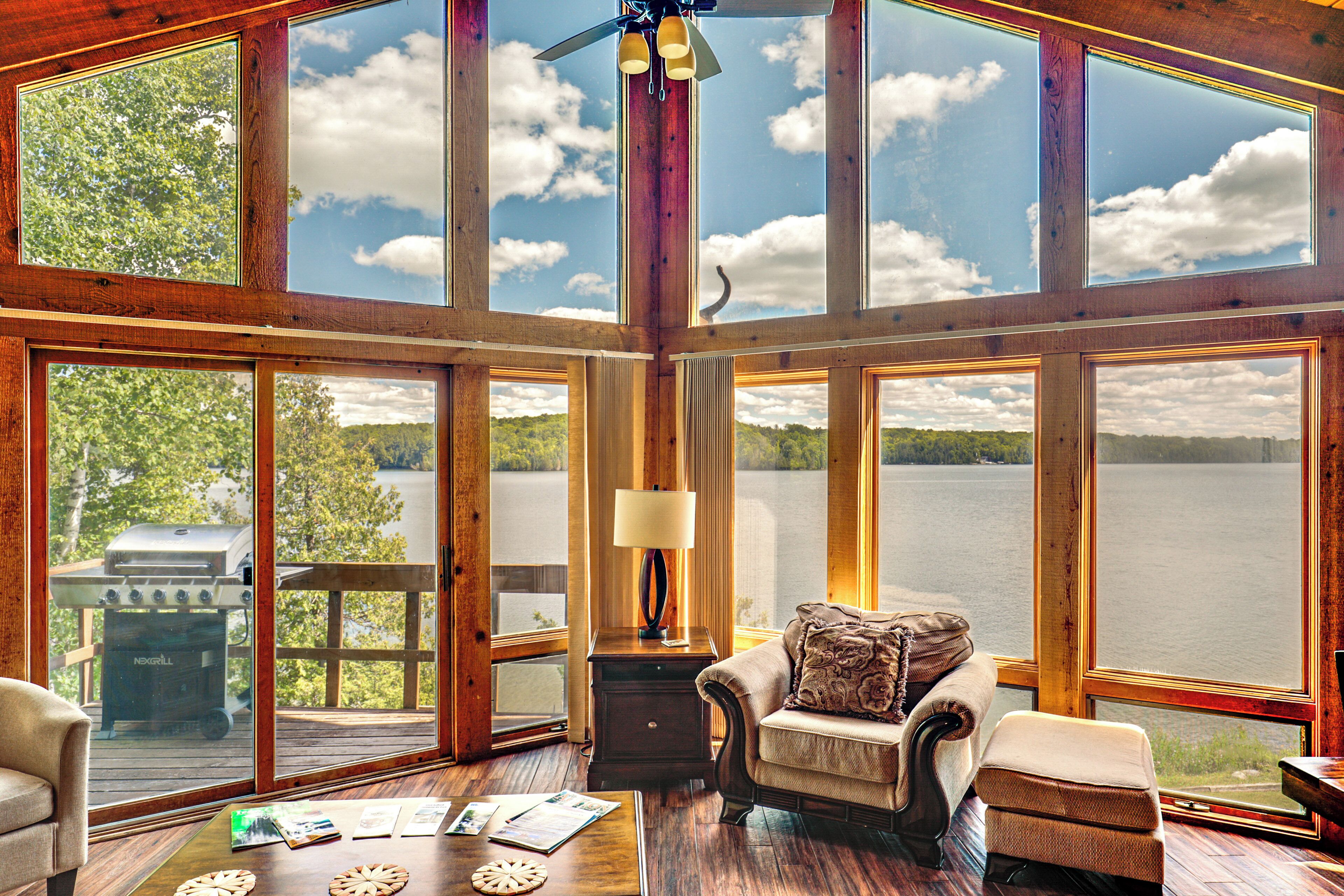 A spacious room with large windows offering a view of a lake and forest. There is a wooden deck outside, a grill, and a cozy seating area with a lamp.
