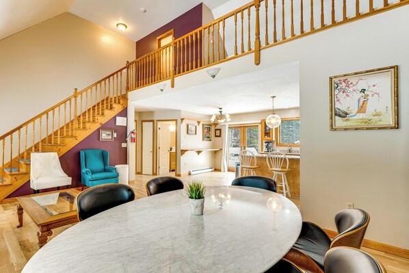 A spacious living area with a round marble table, comfortable seating, a staircase, and a kitchen in the background.