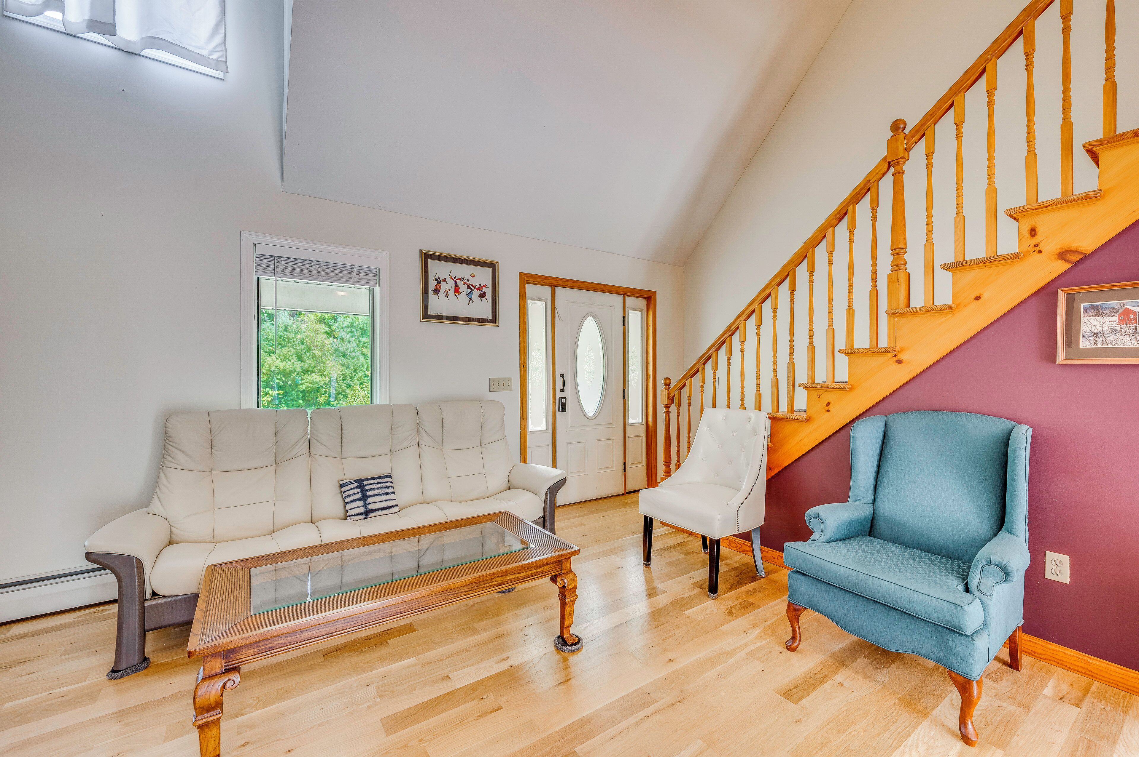 A spacious living room with a wooden staircase, a white sofa, a blue armchair, and a glass coffee table.