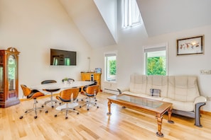 A spacious living area with a high ceiling, a large window, a white sofa, a dining table with chairs, and a wooden coffee table.